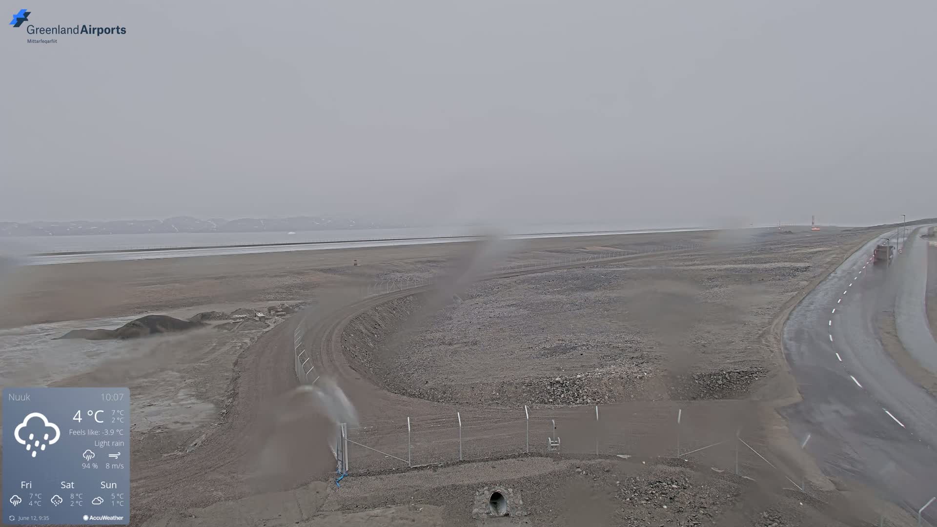 A wide, overcast view shows a flat, gray landscape with a road curving past a fenced-in area under light rain.
