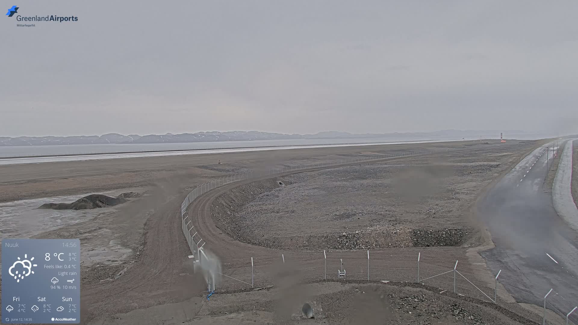 A wide, flat, gray landscape under a light rain with a body of water and distant mountains in the background.