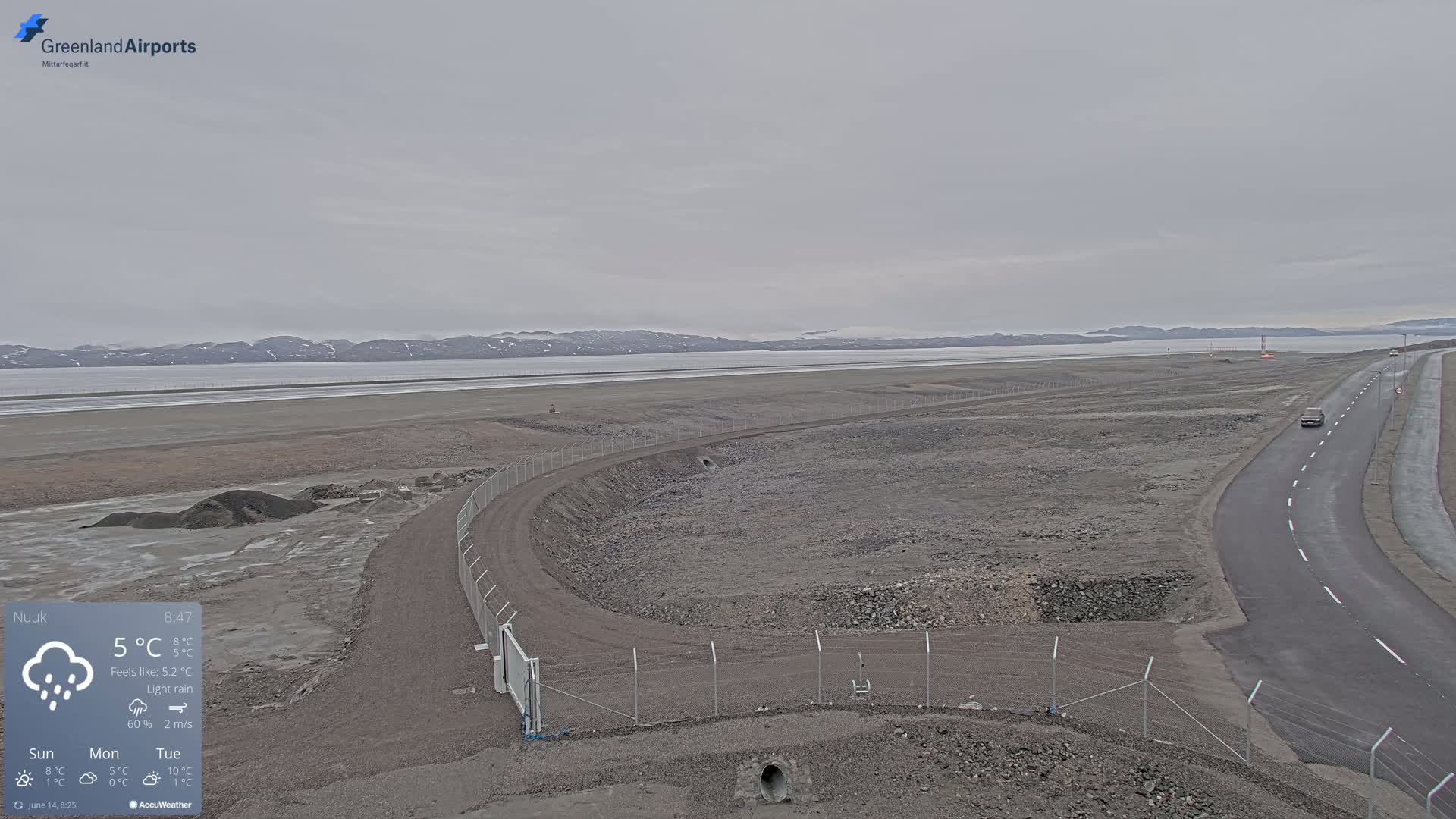 A flat, gray landscape with a paved road and a fenced-in area under a light rain, featuring a body of water and distant snow-capped mountains in the background.