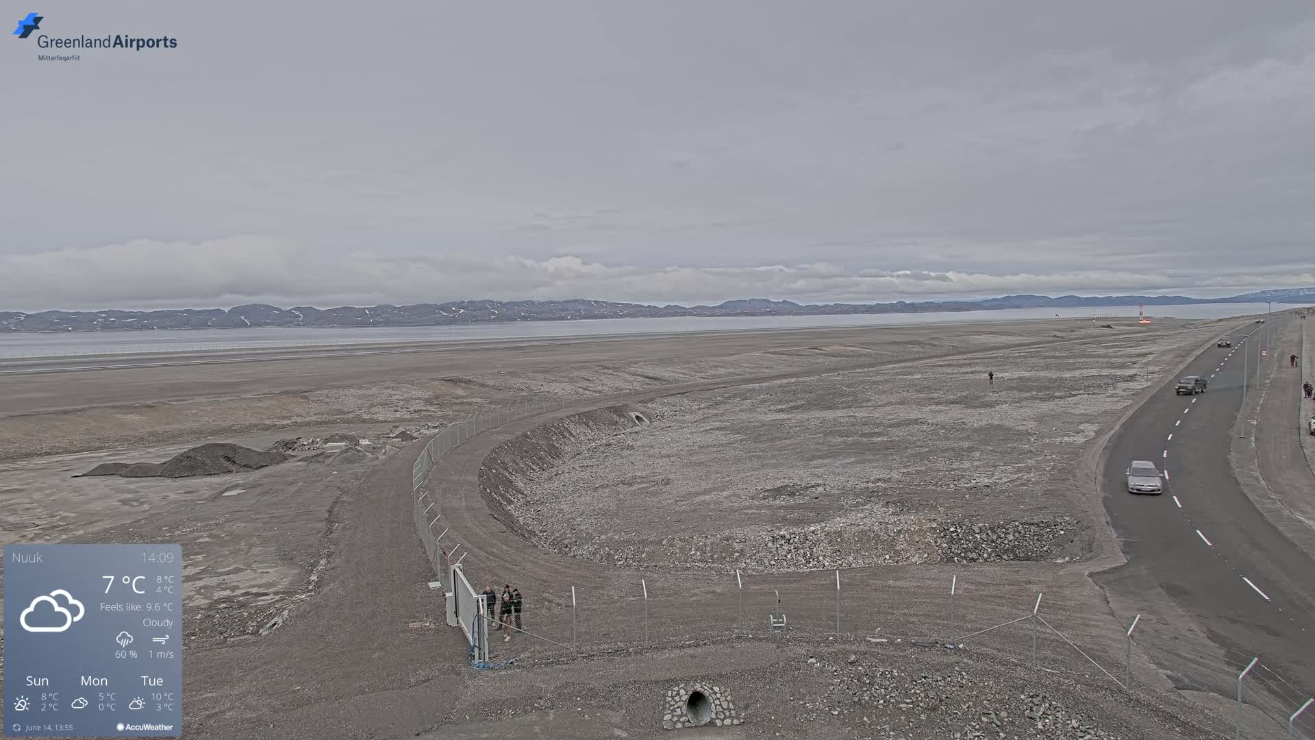 A cloudy, overcast day shows a flat, rocky landscape with a road, a fenced-in area, and a body of water in the background.