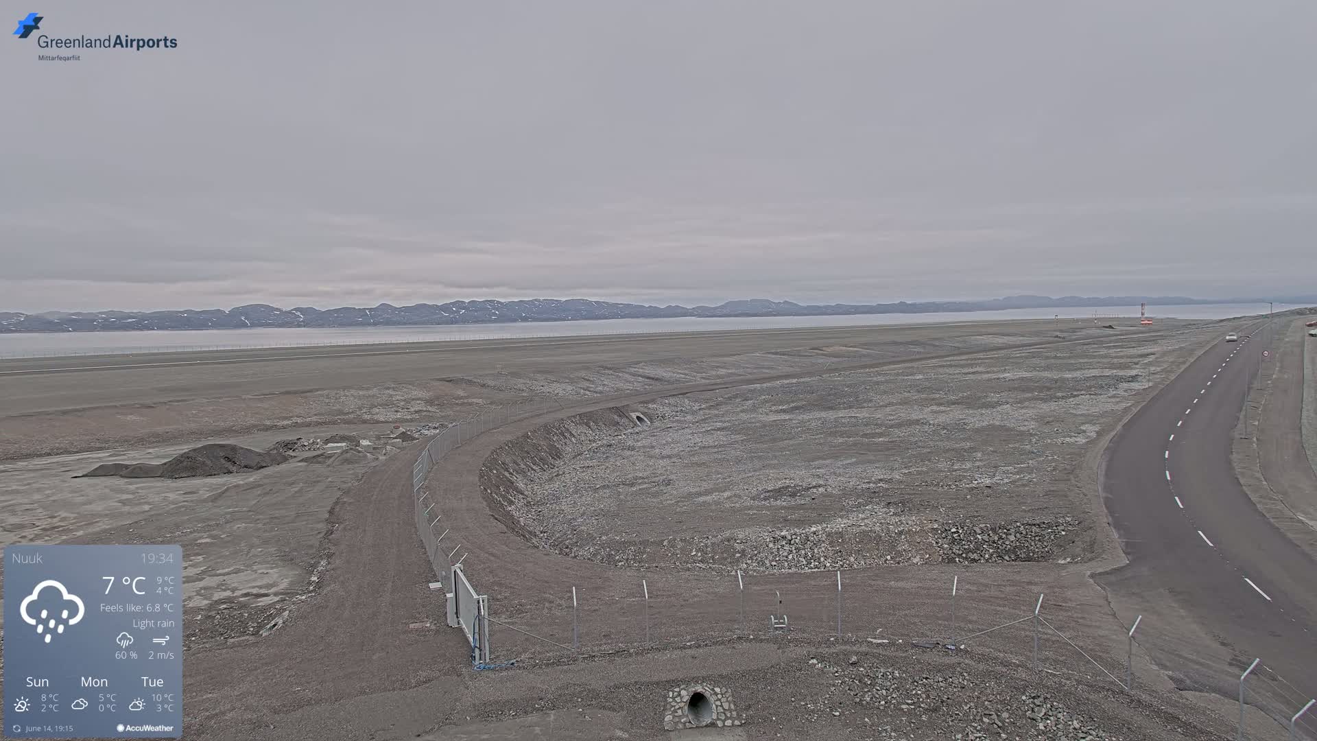 A cloudy, overcast day shows a flat, rocky landscape with a road, a fenced-in area, and a body of water in the background.