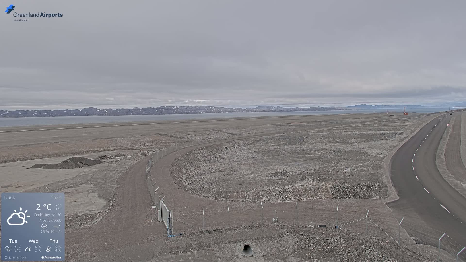 A mostly cloudy day reveals a flat, rocky landscape with a road and a fenced-in area near a body of water and distant mountains.