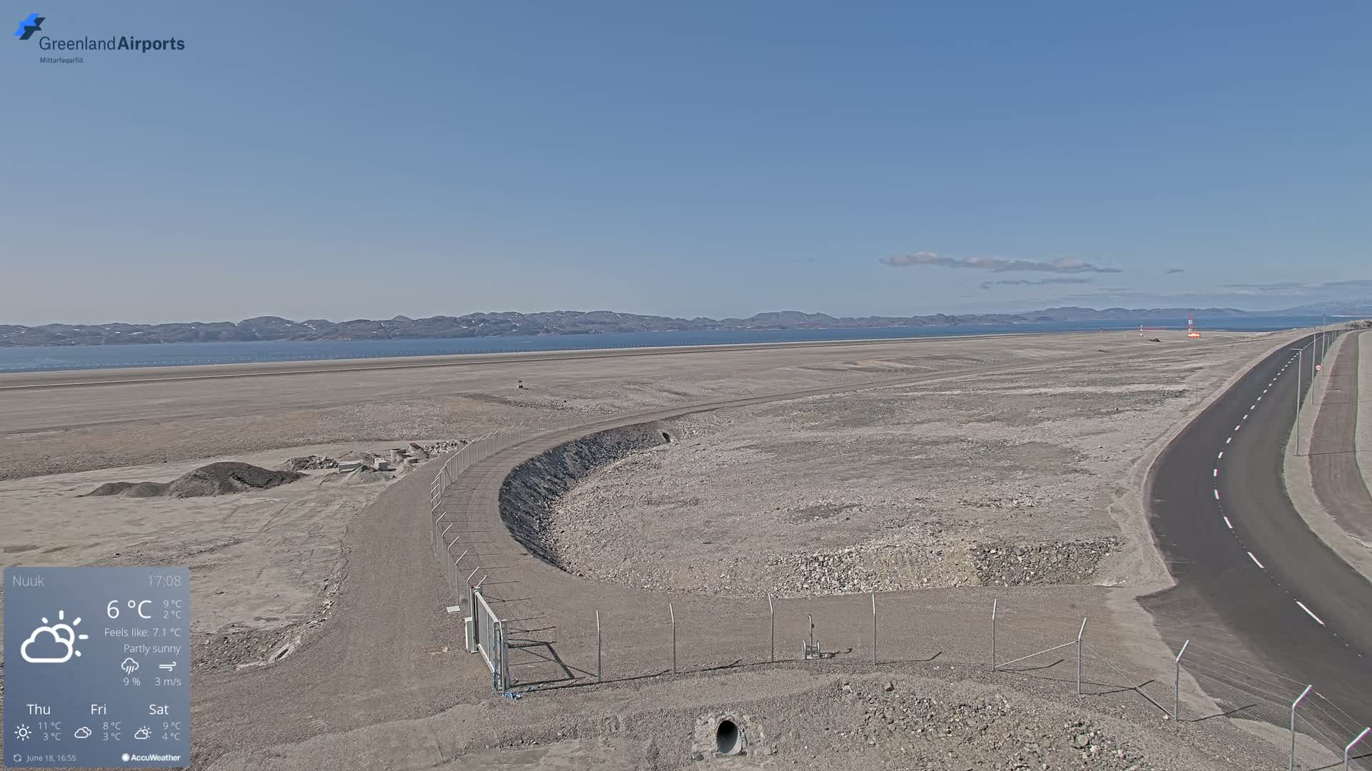A partly sunny, expansive airport tarmac with a paved road curving alongside a fenced-in area, overlooking a calm body of water and distant mountains.
