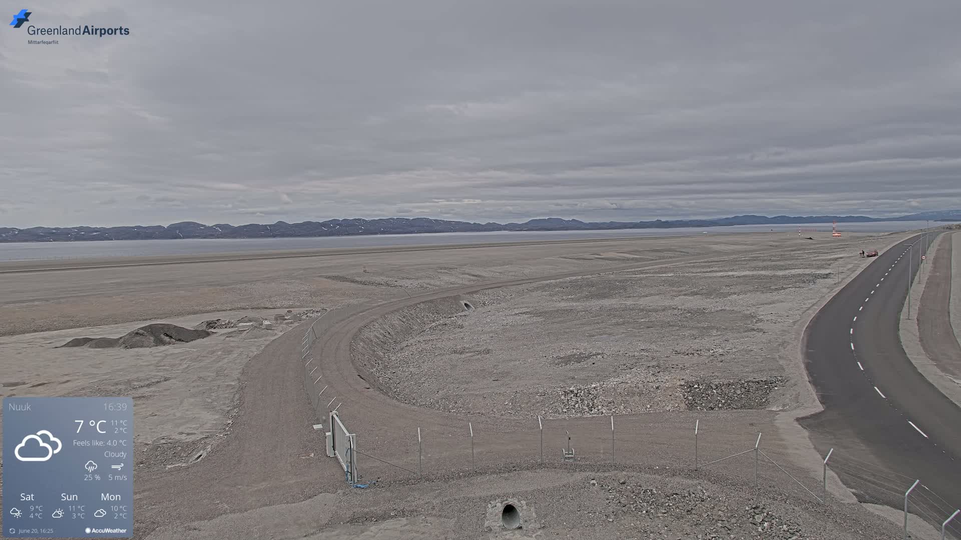 A cloudy, overcast day reveals a flat, rocky landscape with a road and a fenced-in area near a body of water and distant mountains.