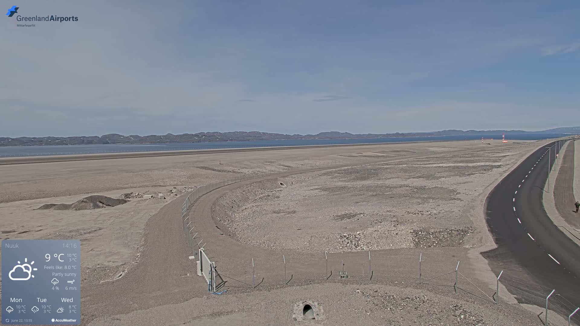 A partly sunny, flat, rocky landscape with a road and airport runway, overlooking a calm body of water and distant mountains.