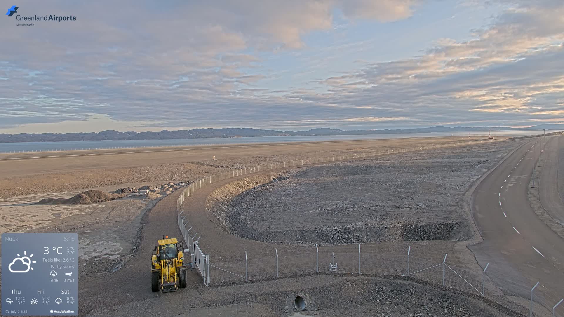 A partly sunny, 3°C day shows a large, flat, barren expanse of land with a road, a fenced-in area undergoing construction, and a yellow piece of construction equipment near a gate, all overlooking a calm body of water and distant mountains.