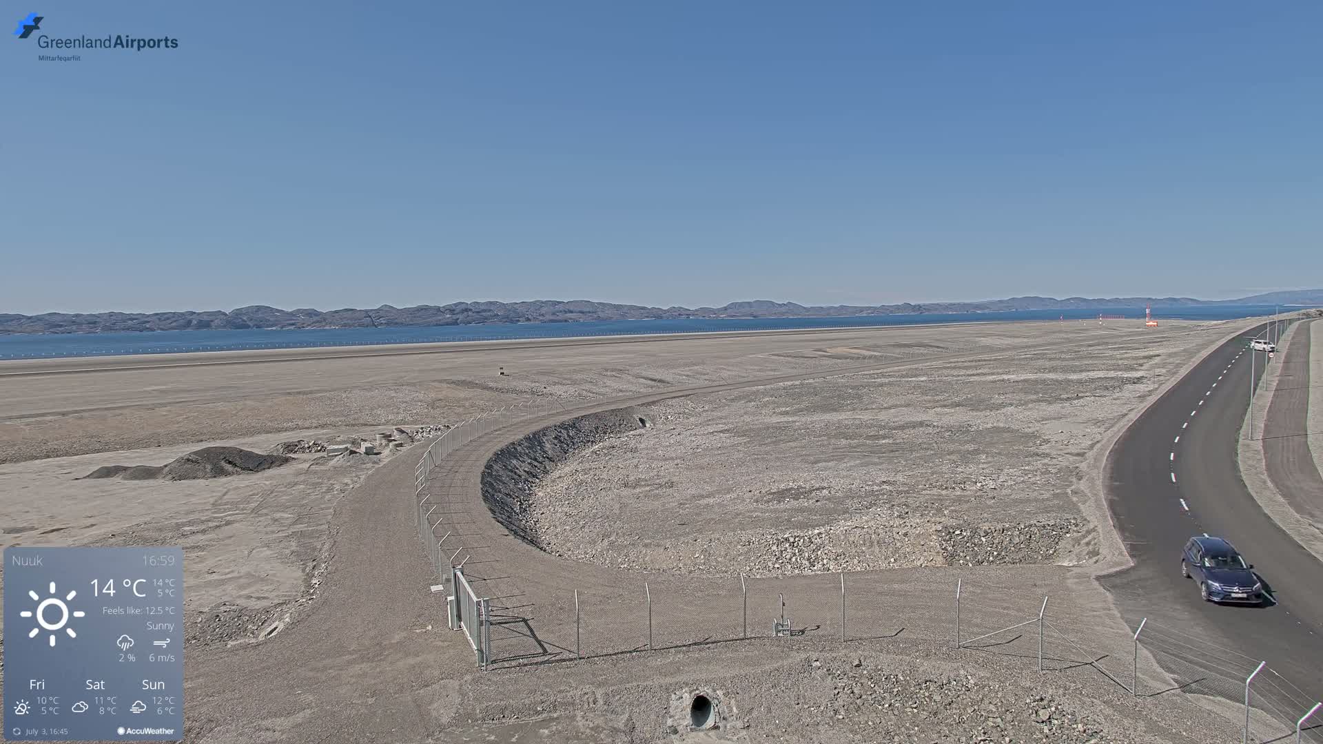 A partly sunny, 3°C day shows a large, flat, barren expanse of land with a road, a fenced-in area undergoing construction, and a yellow piece of construction equipment near a gate, all overlooking a calm body of water and distant mountains.