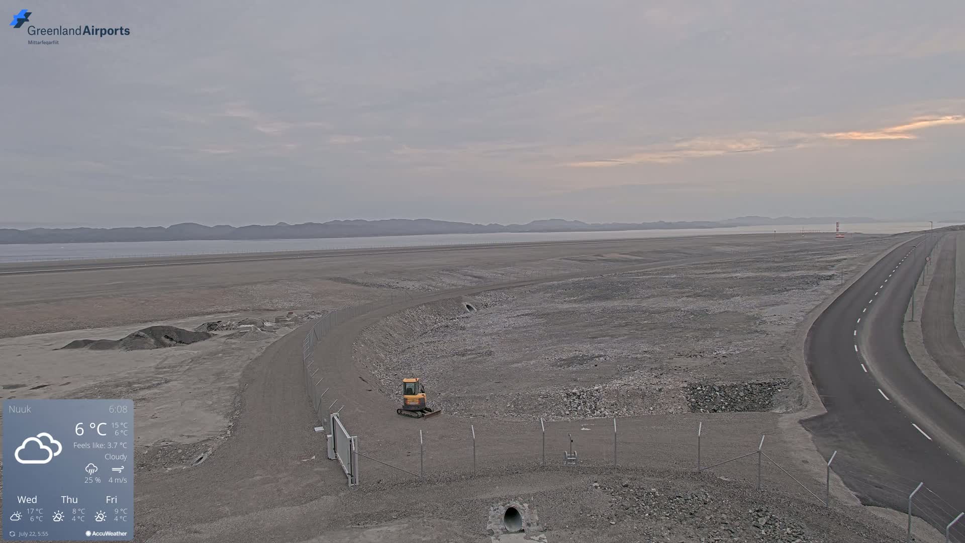 A cloudy day shows a flat, rocky landscape with a small yellow excavator near a fenced-in area, a road curving towards the horizon, and a body of water visible in the distance.