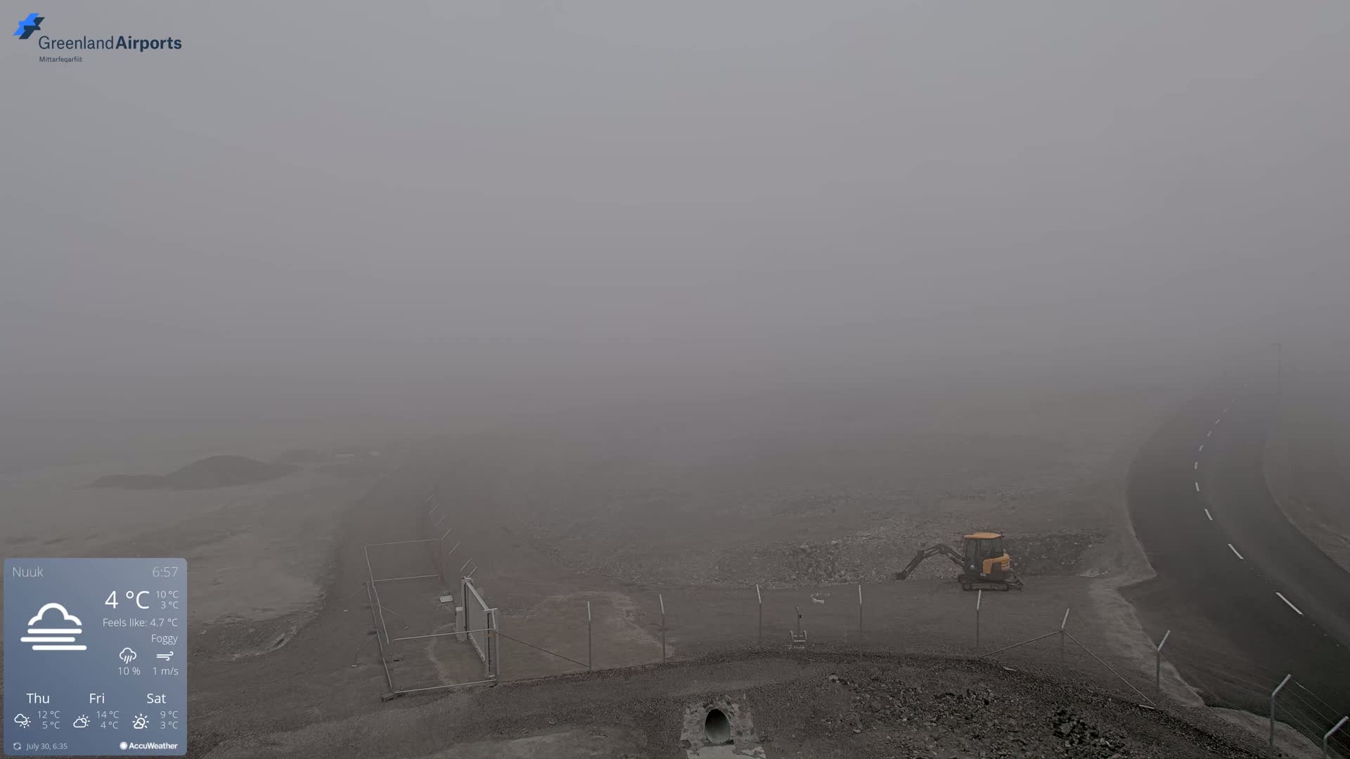 A foggy, overcast landscape shows a small yellow excavator near a fenced-in area next to a road.