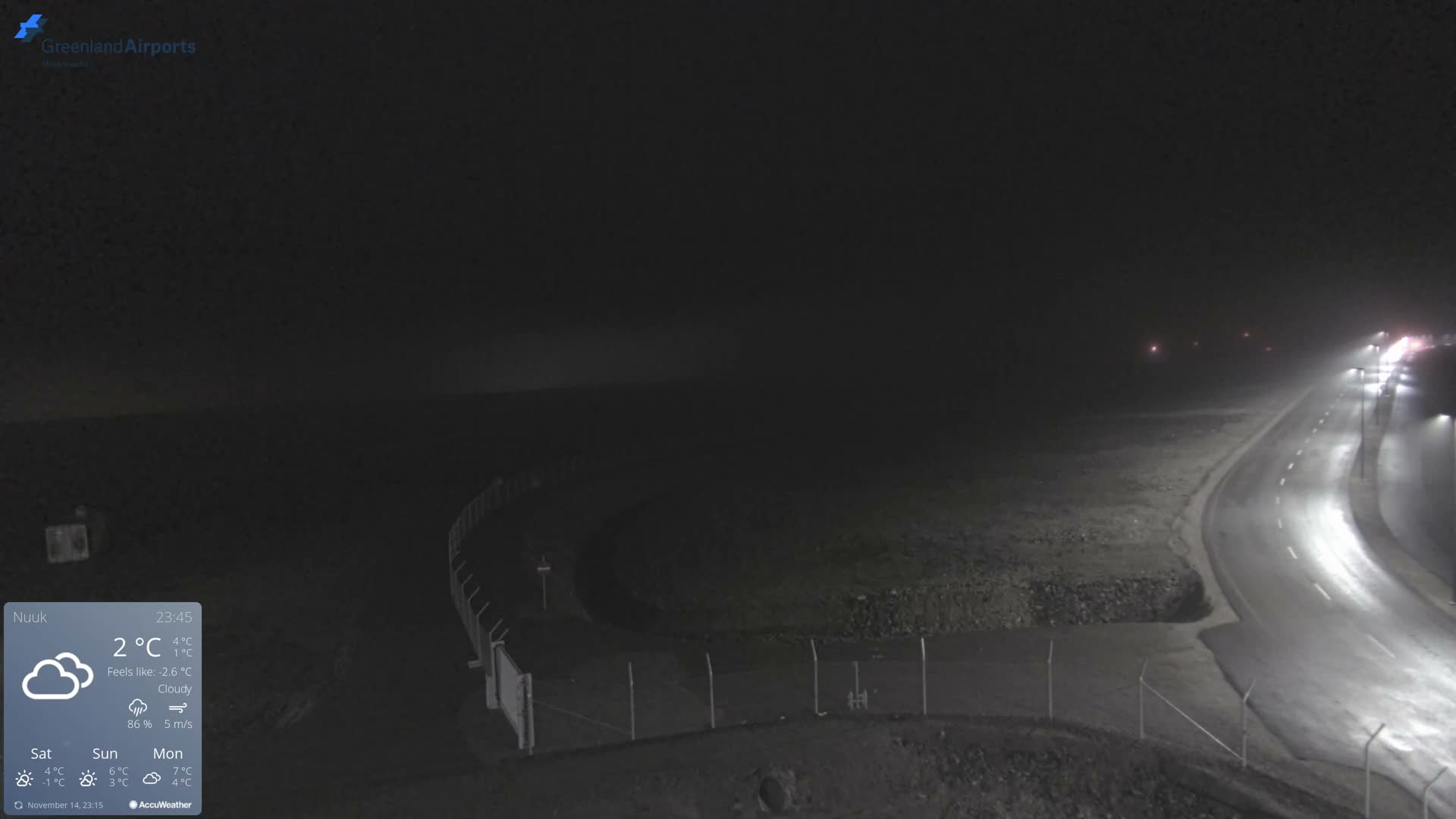 A winding, wet road illuminated by streetlights disappears into heavy fog during a dark night, bordered by a fence and barren ground on the left.