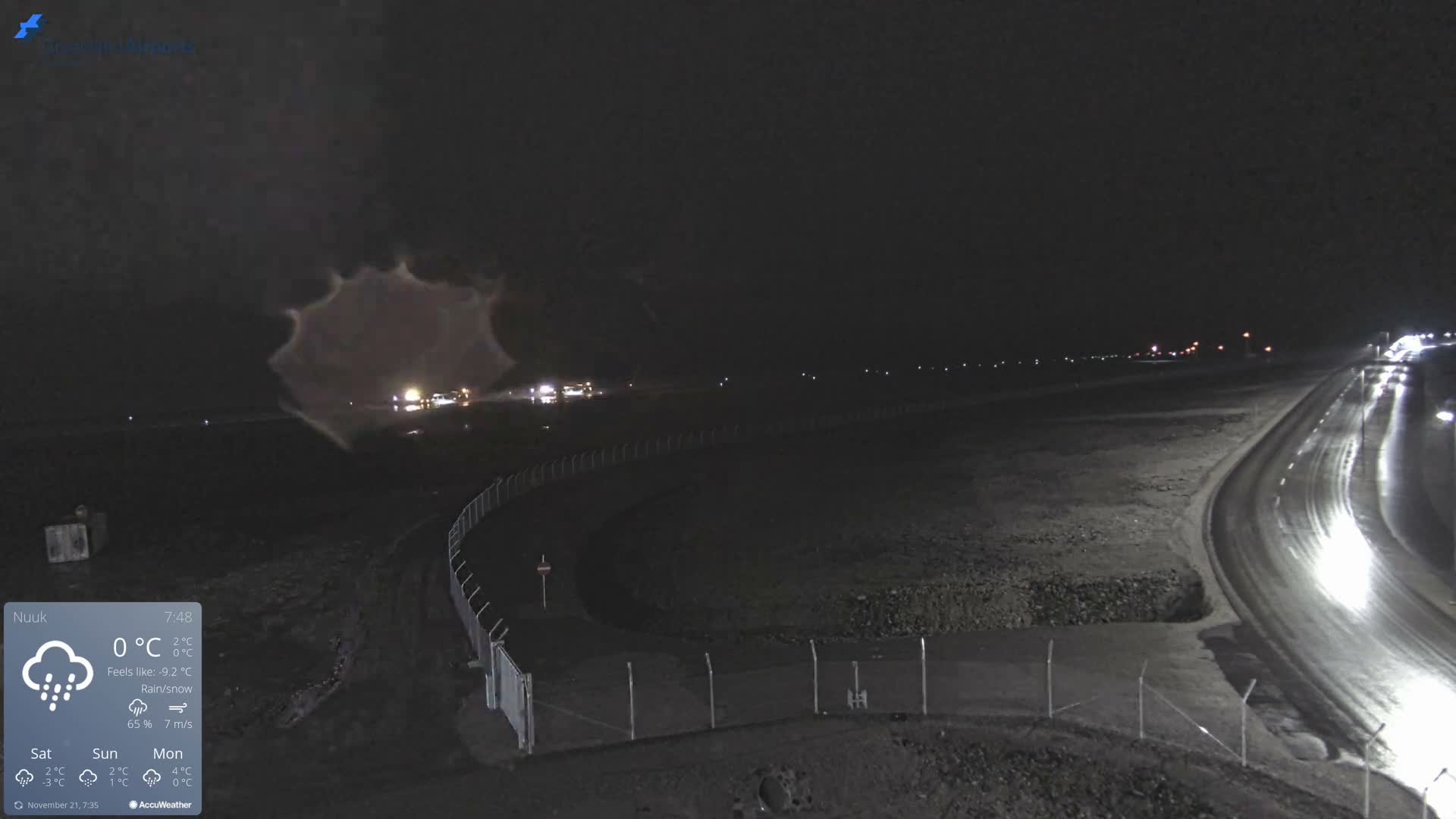 The image depicts a dark, rainy or snowy night outdoors, with a wet and reflective road curving into the distance on the right, bordered by a fence on the left, and distant lights visible on the horizon.