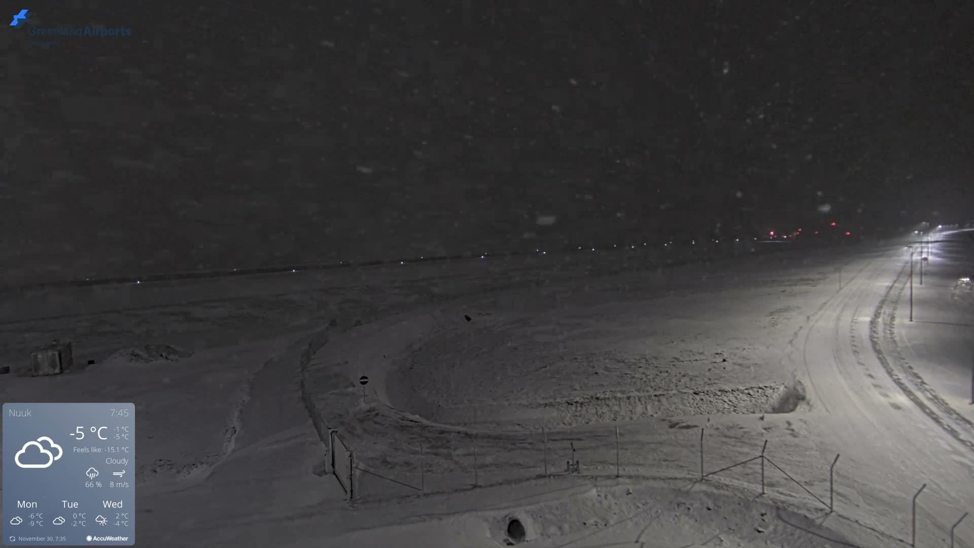 A vast, snow-covered outdoor area is dimly lit at night by streetlights and distant runway lights, with light snow visibly falling from a dark sky.