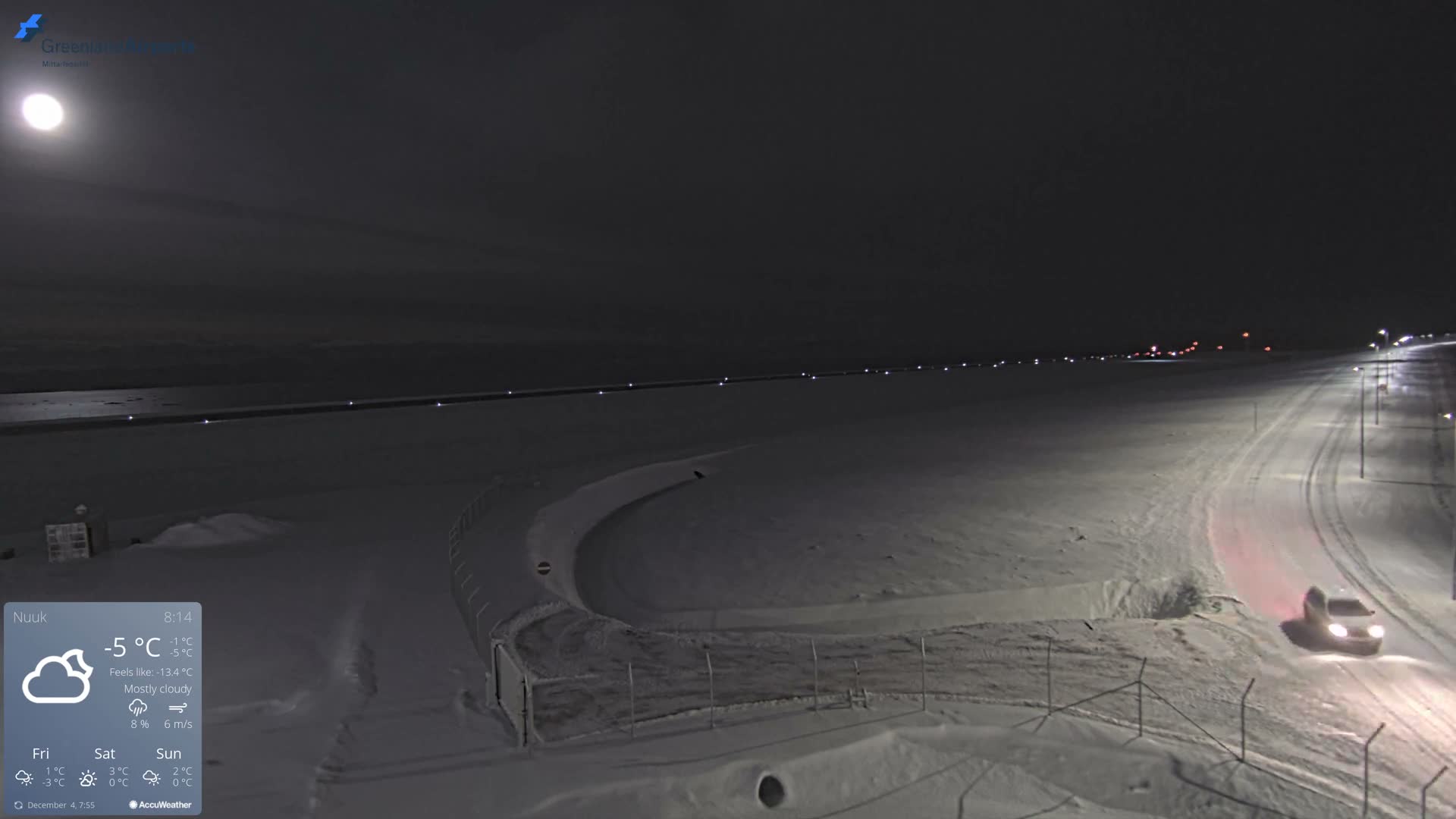 A wide-angle view shows a snow-covered landscape at night under a clear, cold sky, with a bright moon, a long line of distant lights, and a vehicle driving on a streetlight-illuminated, winding snowy road.