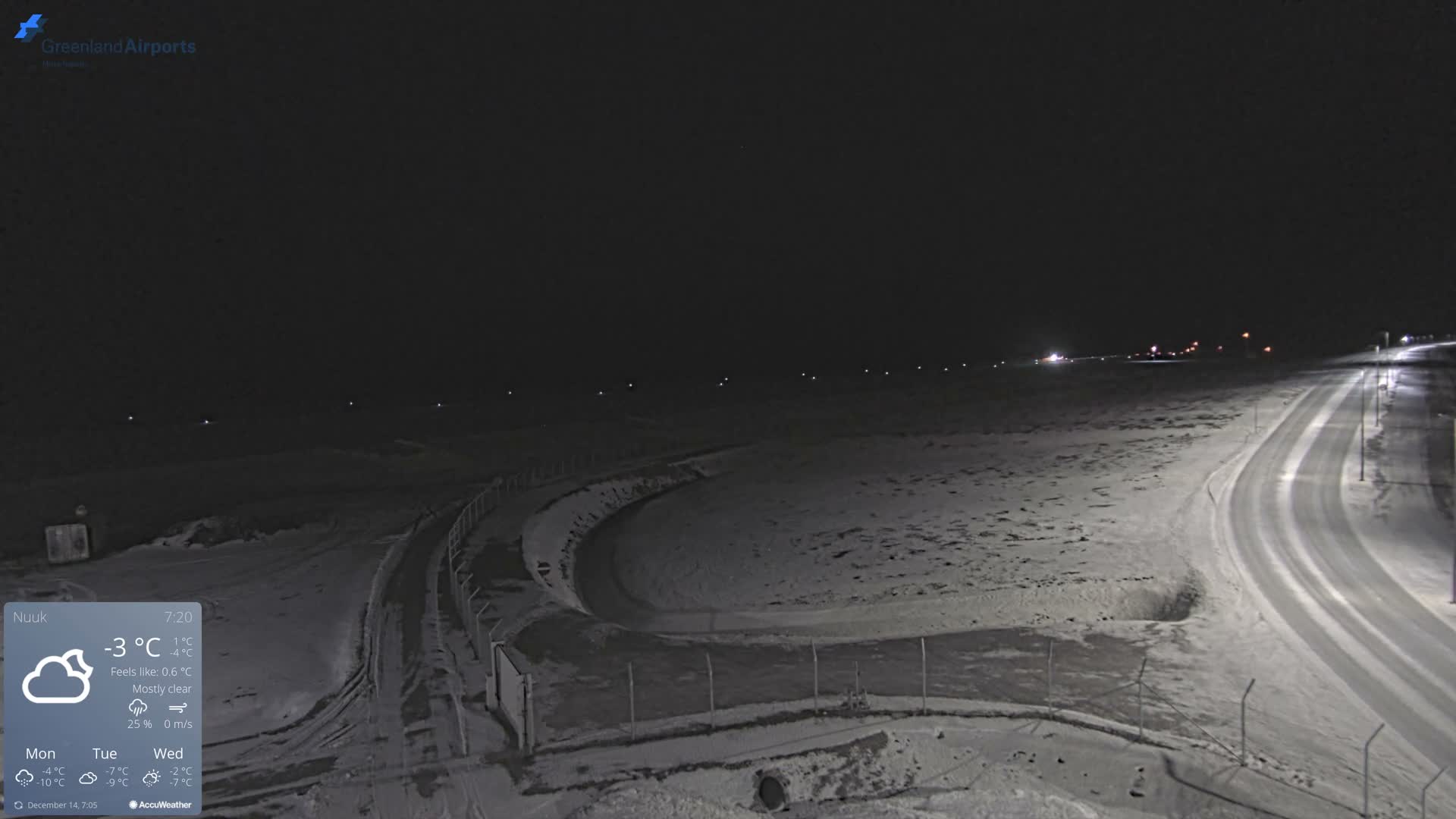 A wide-angle view shows a snow-covered landscape at night under a clear, cold sky, with a bright moon, a long line of distant lights, and a vehicle driving on a streetlight-illuminated, winding snowy road.