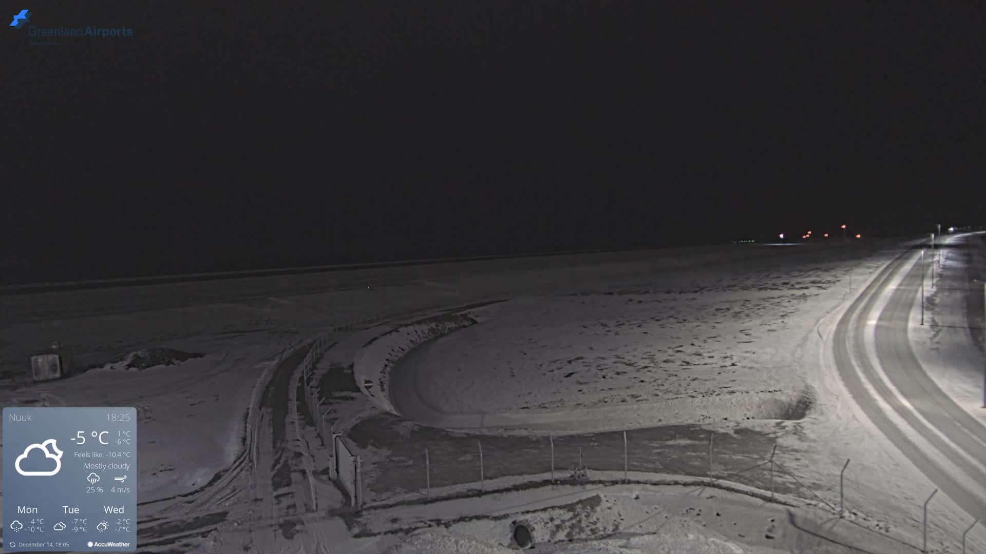 A wide-angle view shows a snow-covered landscape at night under a clear, cold sky, with a bright moon, a long line of distant lights, and a vehicle driving on a streetlight-illuminated, winding snowy road.
