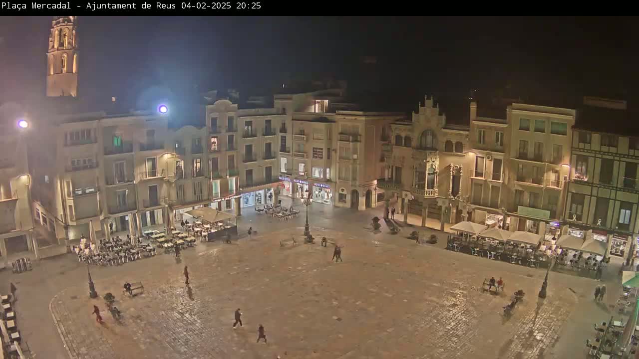 A town square at night, illuminated by streetlights, with several people and outdoor seating areas around its perimeter.
