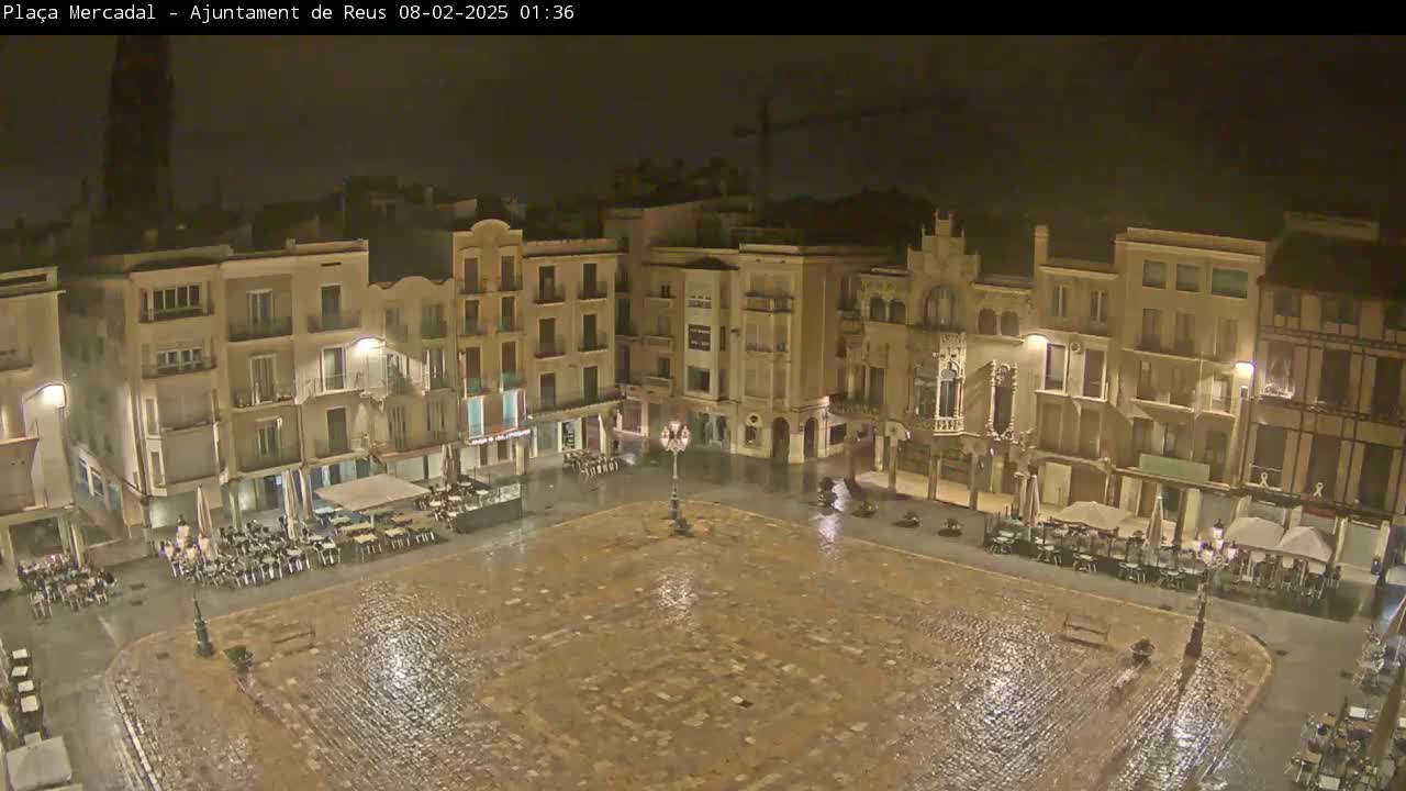 A rain-soaked town square at night, surrounded by illuminated buildings with outdoor seating areas.