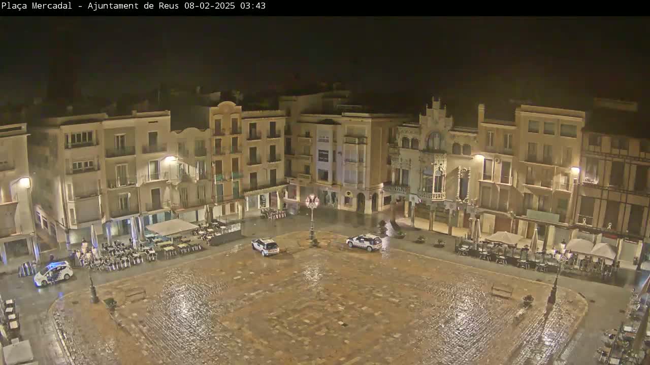 A rain-slicked, cobblestone town square at night is surrounded by multi-story buildings with outdoor seating areas, and two police cars are parked in the center.