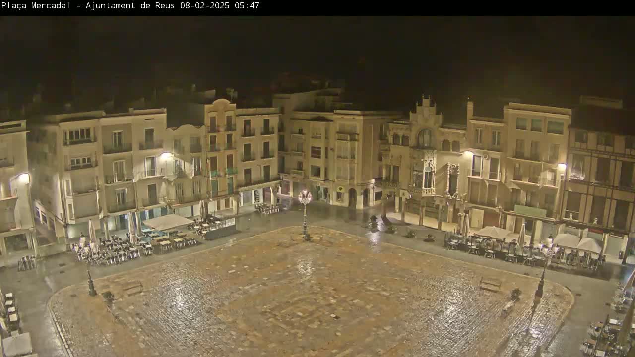 A rain-slicked, cobblestone town square at night, surrounded by multi-story buildings with outdoor seating areas under umbrellas.