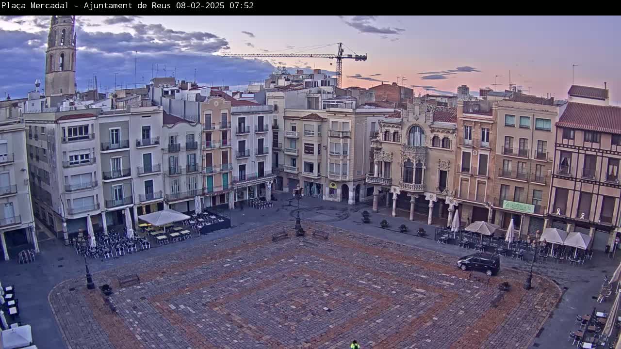 A large, cobblestone town square with several multi-story buildings surrounding it, under a twilight sky with partly cloudy conditions.