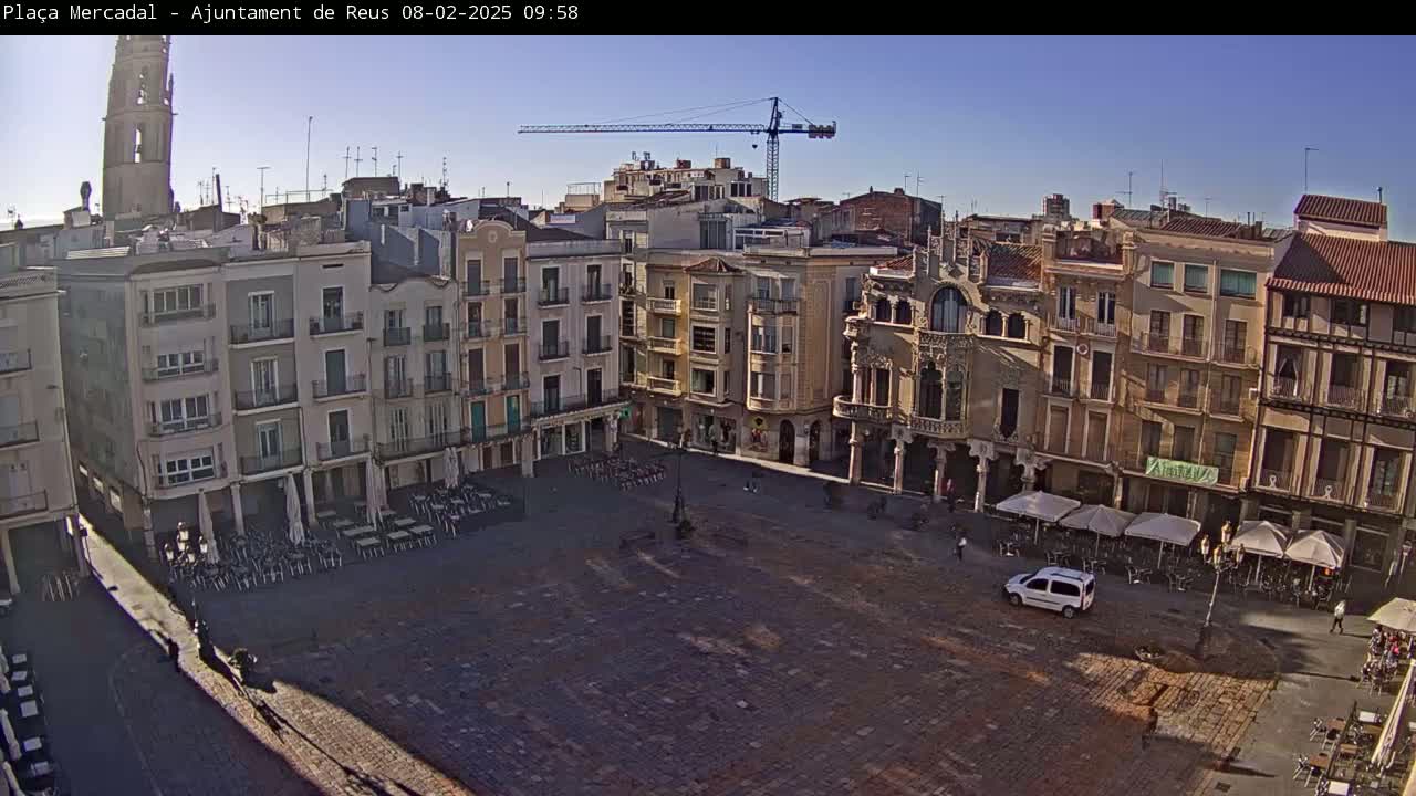A large, mostly empty town square surrounded by multi-story buildings under a clear, sunny sky, with a construction crane visible in the distance.