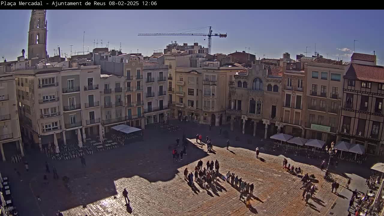 A sunny day reveals a large town square with multiple story buildings surrounding it, several people walking around, and outdoor seating areas with umbrellas.