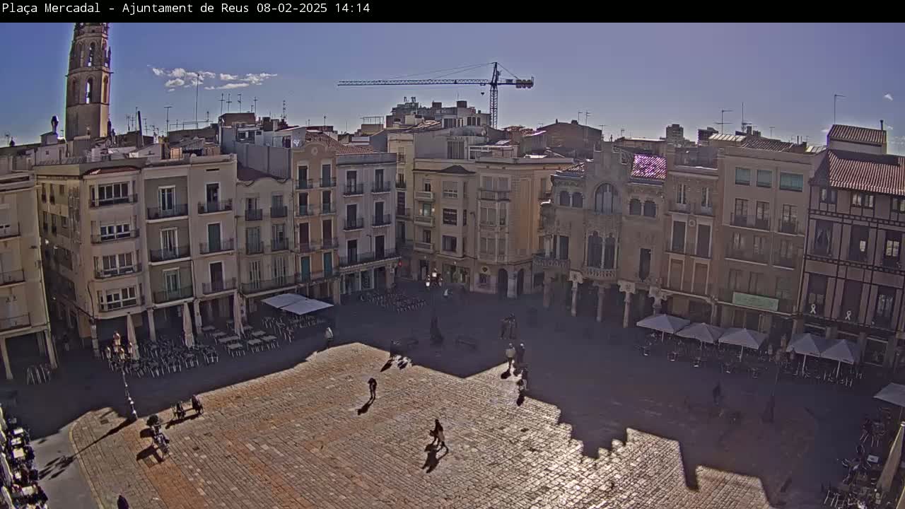 A large, sunlit town square is surrounded by multi-story buildings, with a few people walking around and some outdoor seating areas under umbrellas.