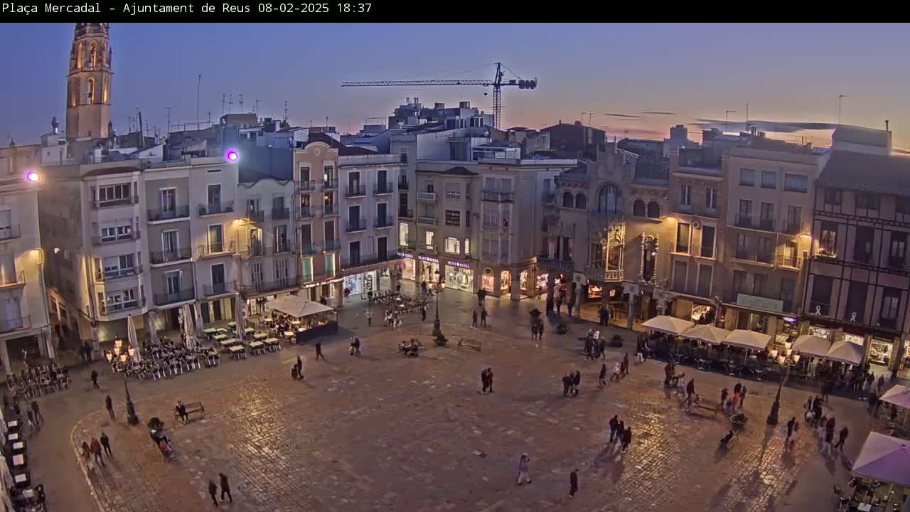 A town square at dusk, with people walking around and sitting at outdoor cafes under dim lighting, is surrounded by multi-story buildings.