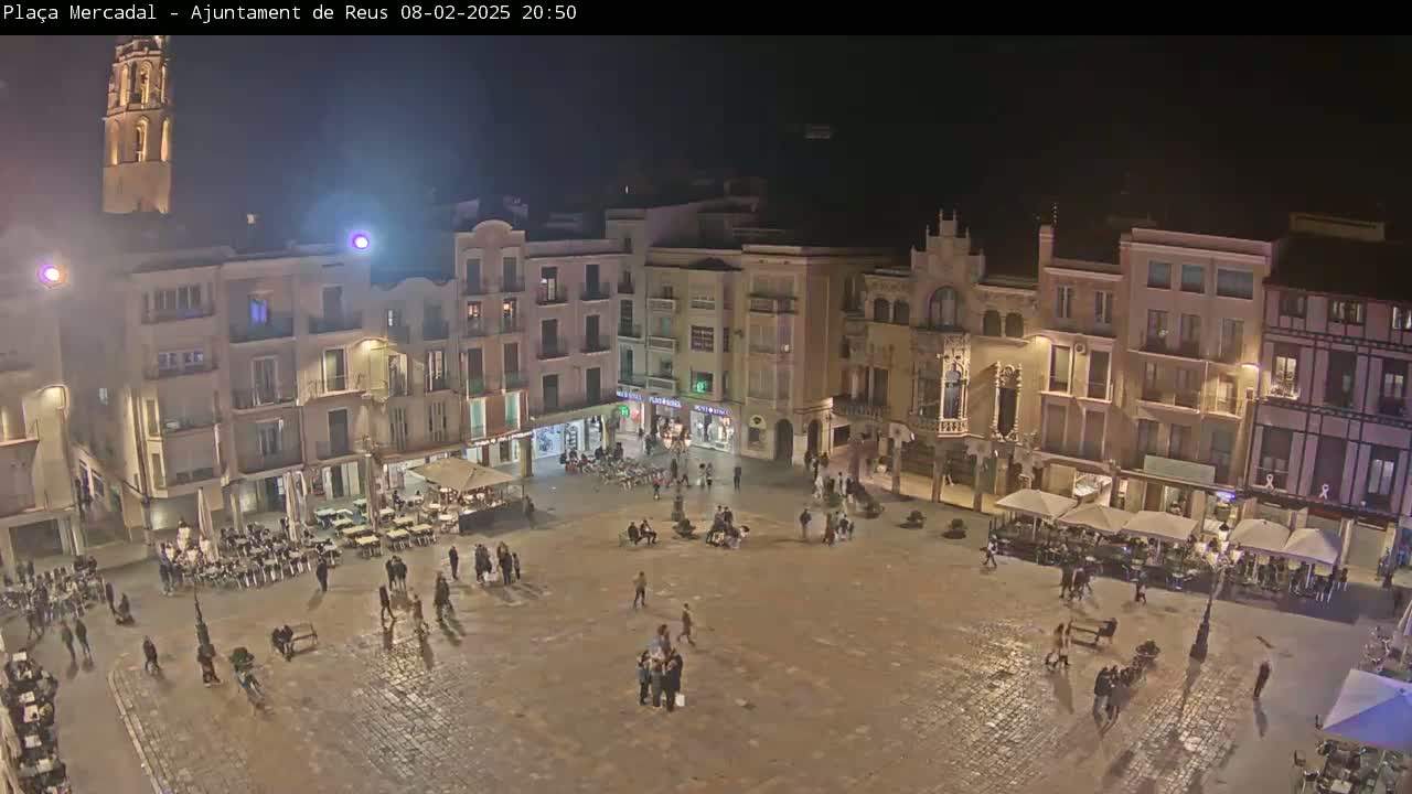 A nighttime view of a town square, with several buildings surrounding it and people gathered in the square under a clear night sky.