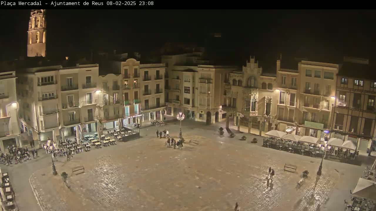 A nighttime, high-angle view of a town square, surrounded by multi-story buildings with outdoor seating areas and a few pedestrians.