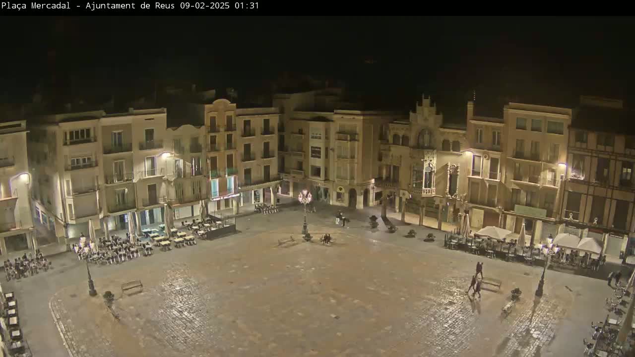 A large, mostly empty town square at night, surrounded by multi-story buildings with outdoor seating areas, is lightly wet from recent rain.