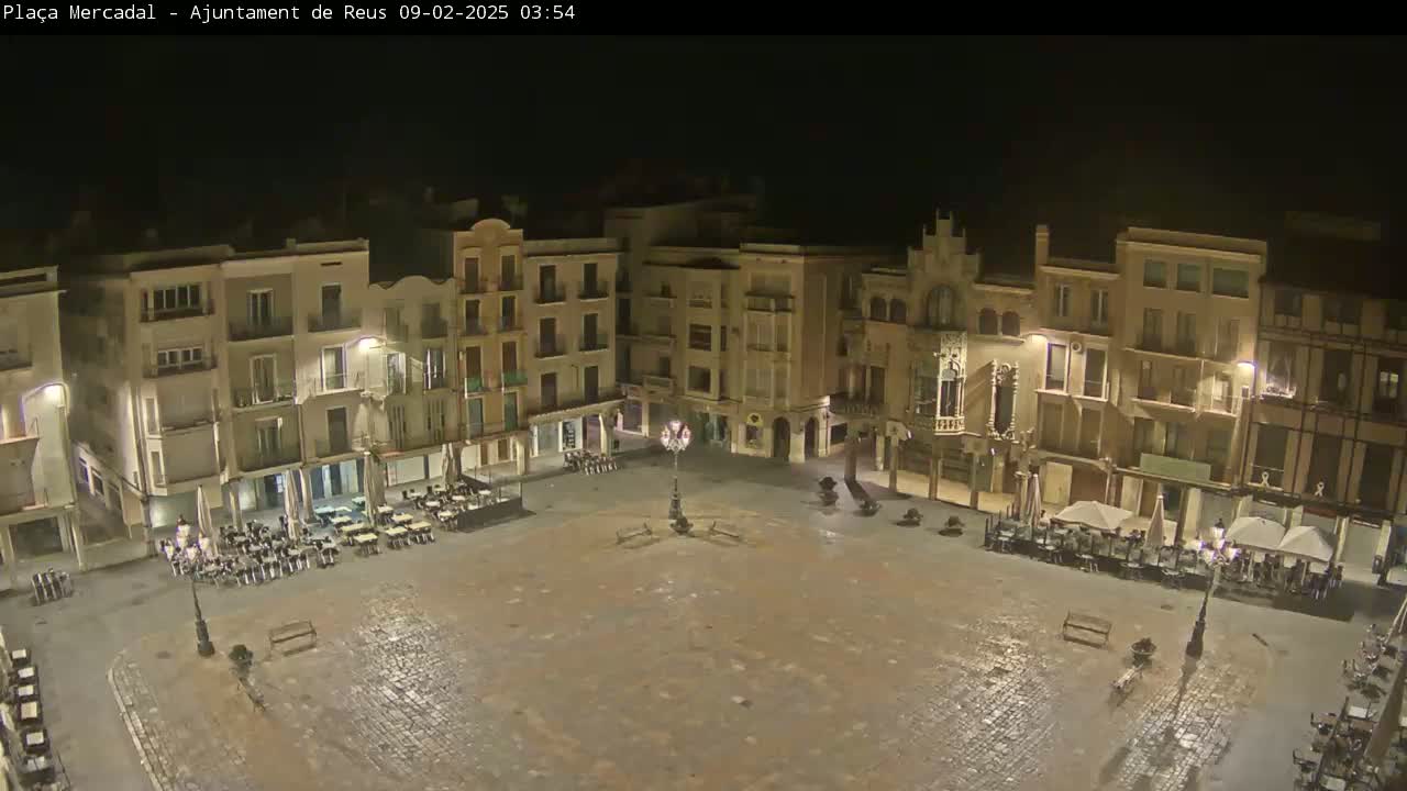 An empty town square at night, surrounded by multi-story buildings with outdoor seating areas, is lightly wet.