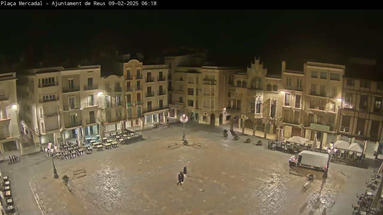 An empty town square at night, surrounded by multi-story buildings with outdoor seating areas, is lightly illuminated.