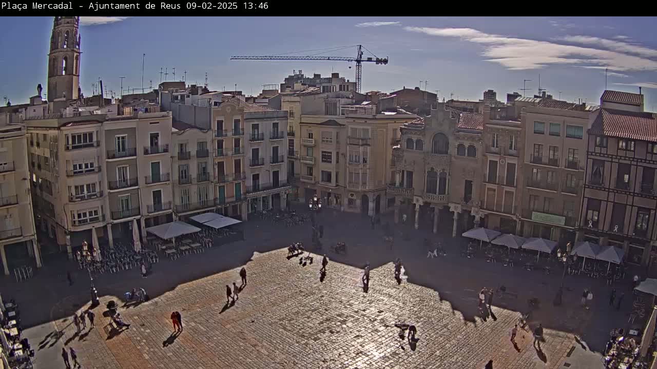 A sunny day reveals a large, cobblestone plaza surrounded by multi-story buildings, with people strolling and some seated at outdoor cafes.