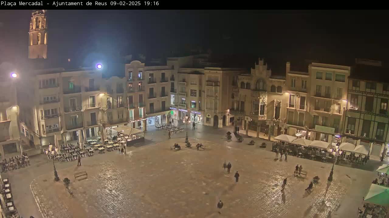 A nighttime, high-angle view of a sparsely populated town square surrounded by multi-story buildings with outdoor seating areas under umbrellas.