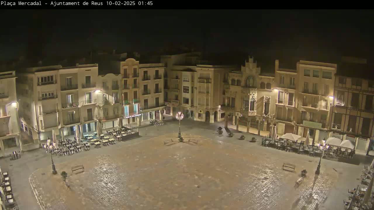 A wet, empty town square at night, surrounded by multi-story buildings with outdoor seating areas.