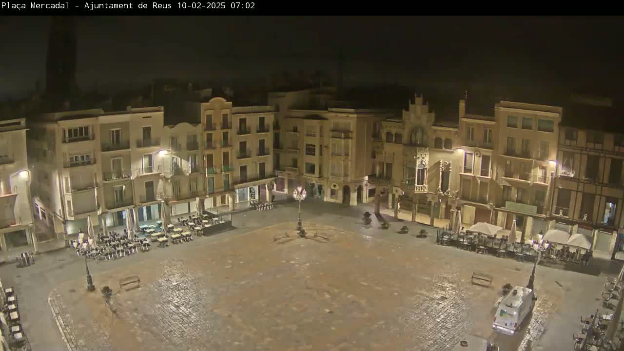A large, mostly empty town square at night, surrounded by multi-story buildings with outdoor seating areas, is lightly wet from a recent rain.