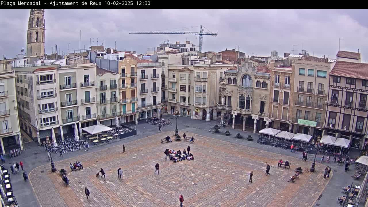 A large, paved town square with several outdoor cafes and a few people is surrounded by multi-story buildings under an overcast sky.