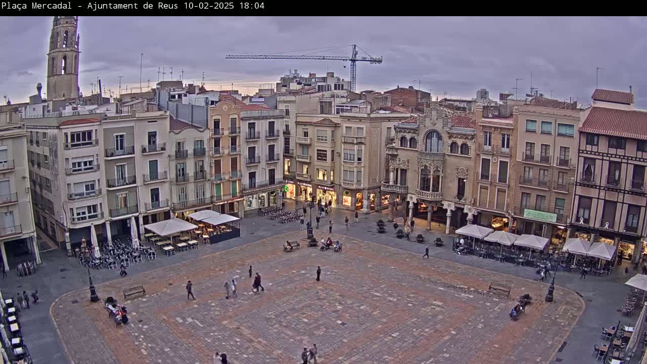 A large, paved town square is surrounded by multi-story buildings under an overcast sky at dusk, with several outdoor cafes set up in the square.