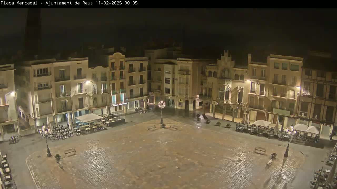 A rain-slicked town square at night, surrounded by illuminated buildings with outdoor seating areas.