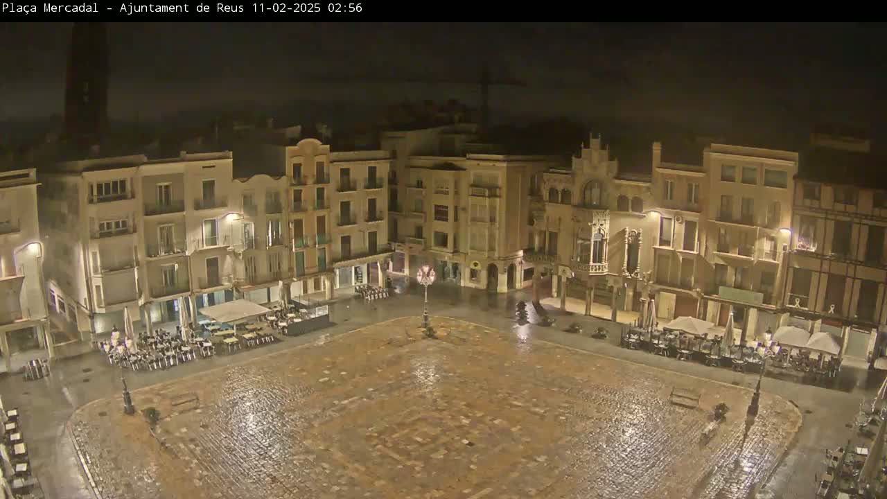 A rain-slicked town square at night, surrounded by multi-story buildings with outdoor seating areas.