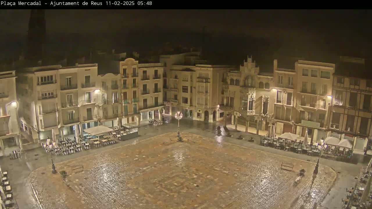 A rain-slicked town square at night, surrounded by illuminated buildings with outdoor seating areas.