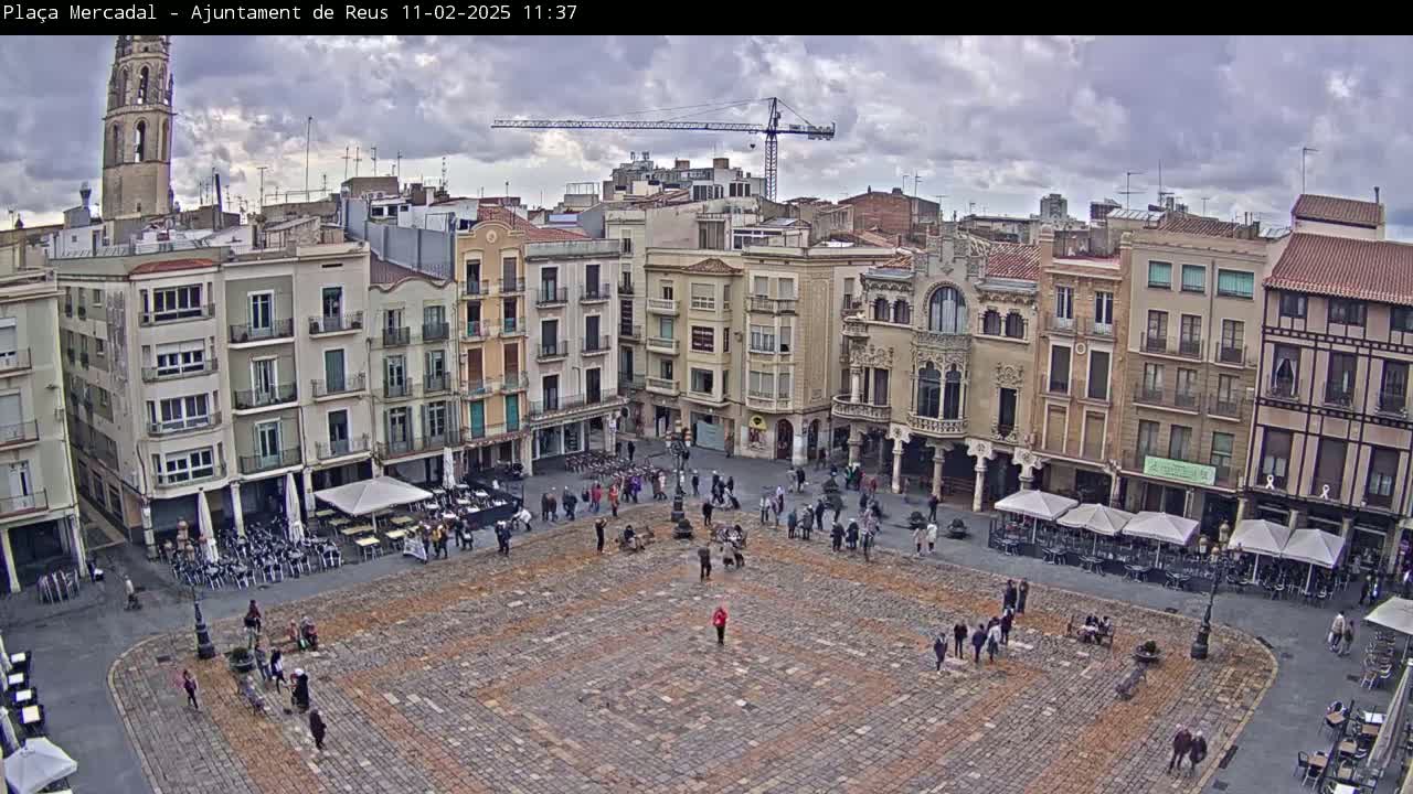 A large town square paved with stone is surrounded by multi-story buildings under a cloudy sky, with a few people walking around and outdoor seating areas in front of some of the buildings.