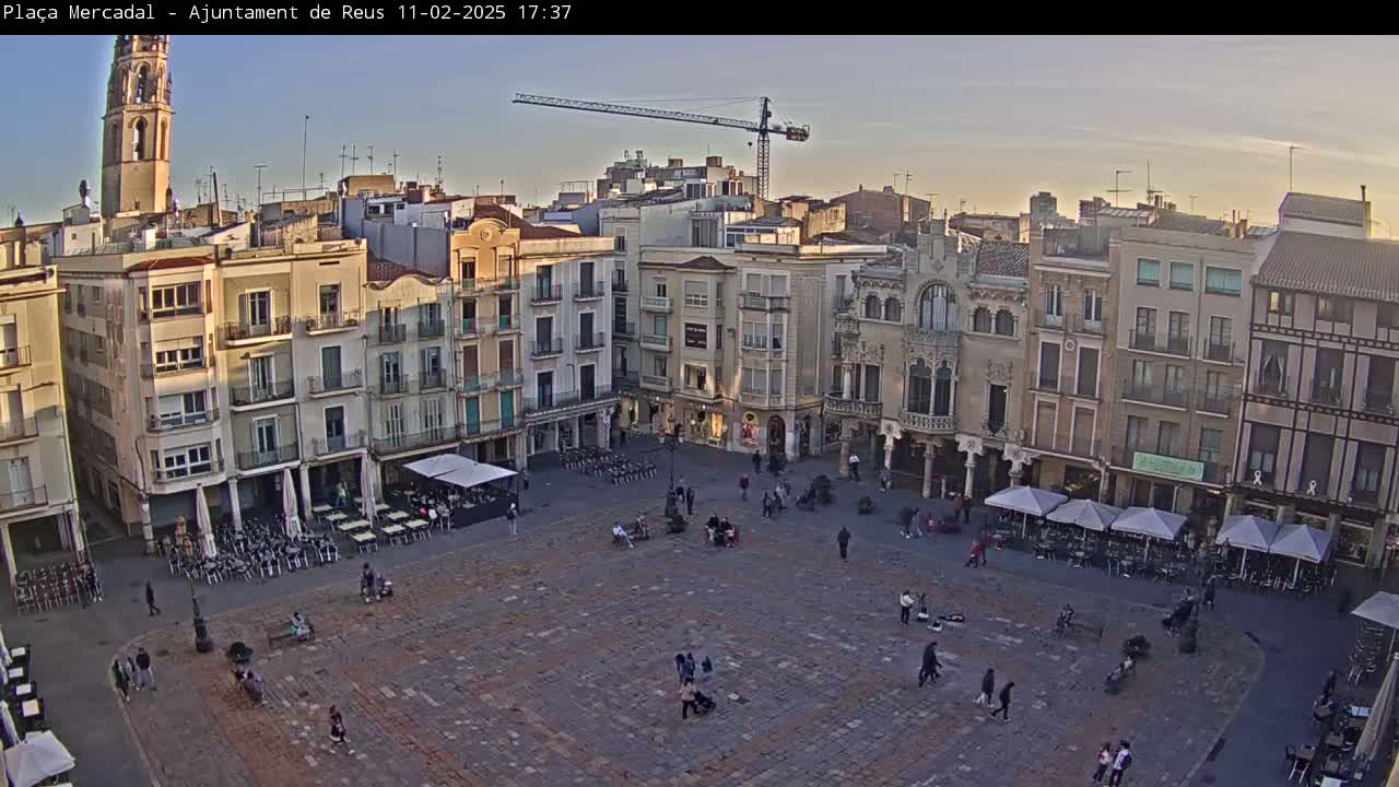 A large town square, paved with stone, is filled with sparsely scattered people and outdoor seating areas under umbrellas, surrounded by multi-story buildings under a clear, twilight sky.
