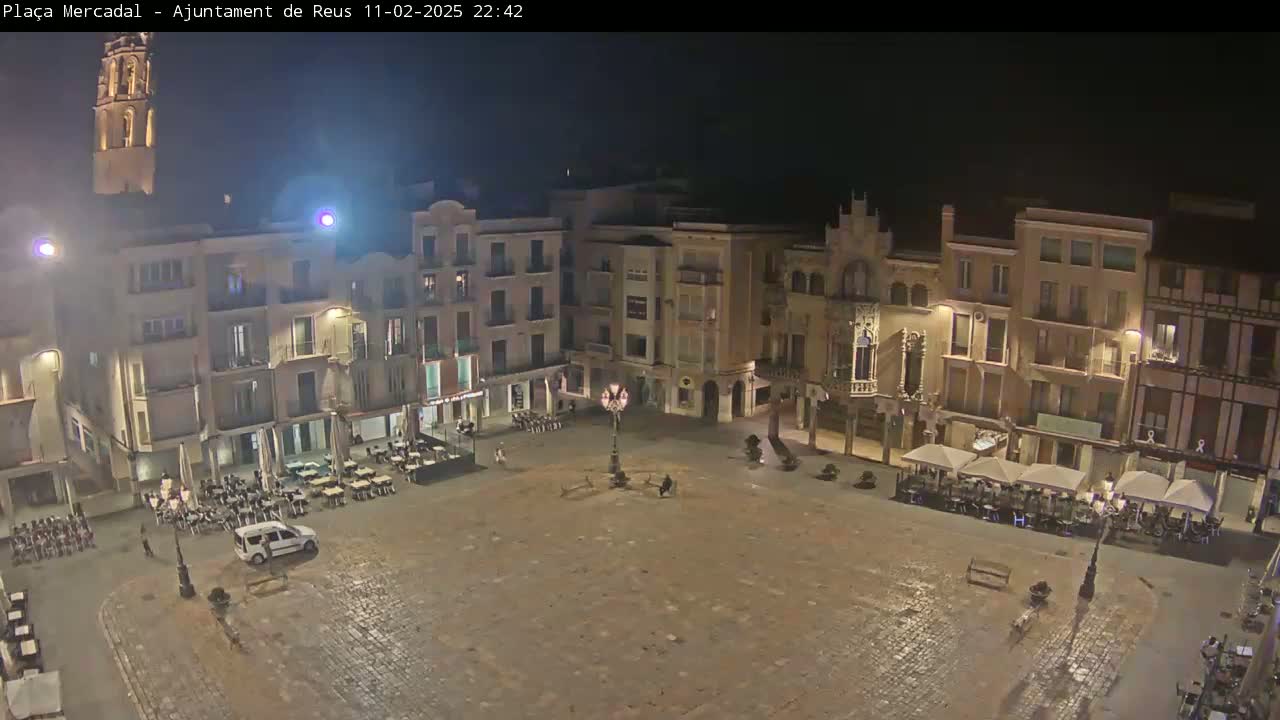 A mostly empty, paved town square at night is surrounded by multi-story buildings with outdoor seating areas.