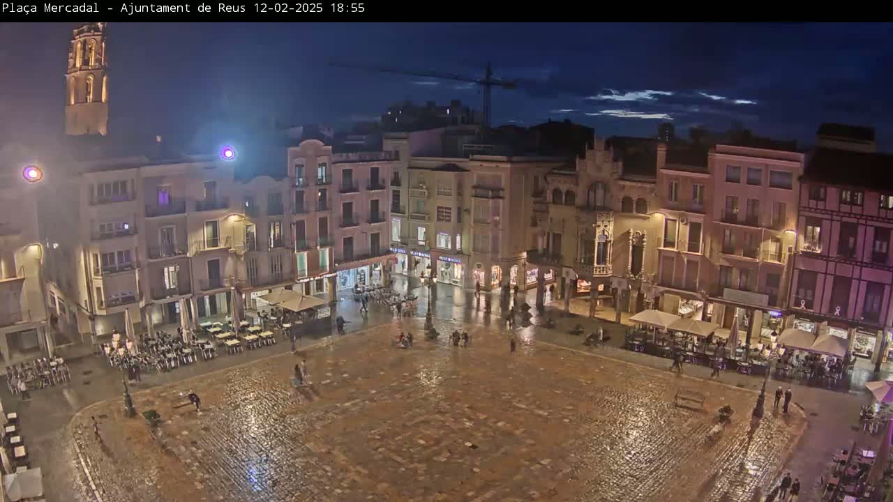 A rain-slicked town square at night, surrounded by illuminated buildings and sparsely populated with pedestrians.