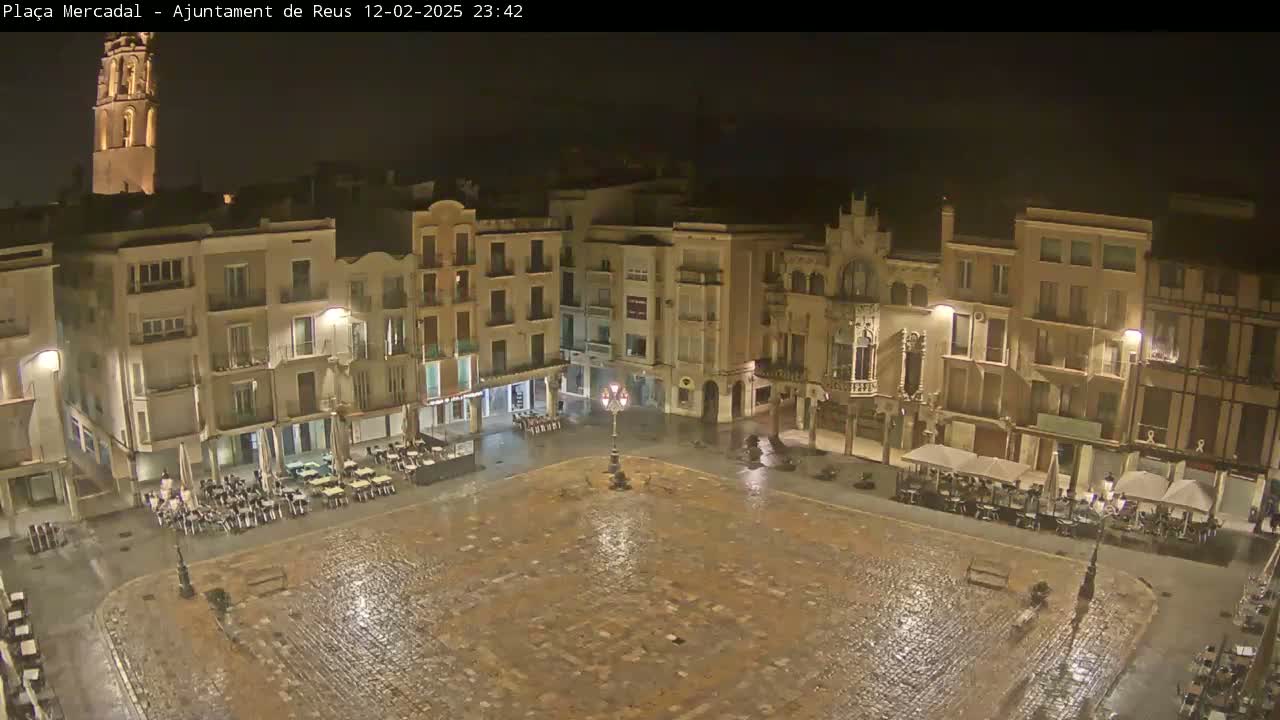 A rain-slicked town square at night, surrounded by multi-story buildings with illuminated windows and outdoor seating areas.