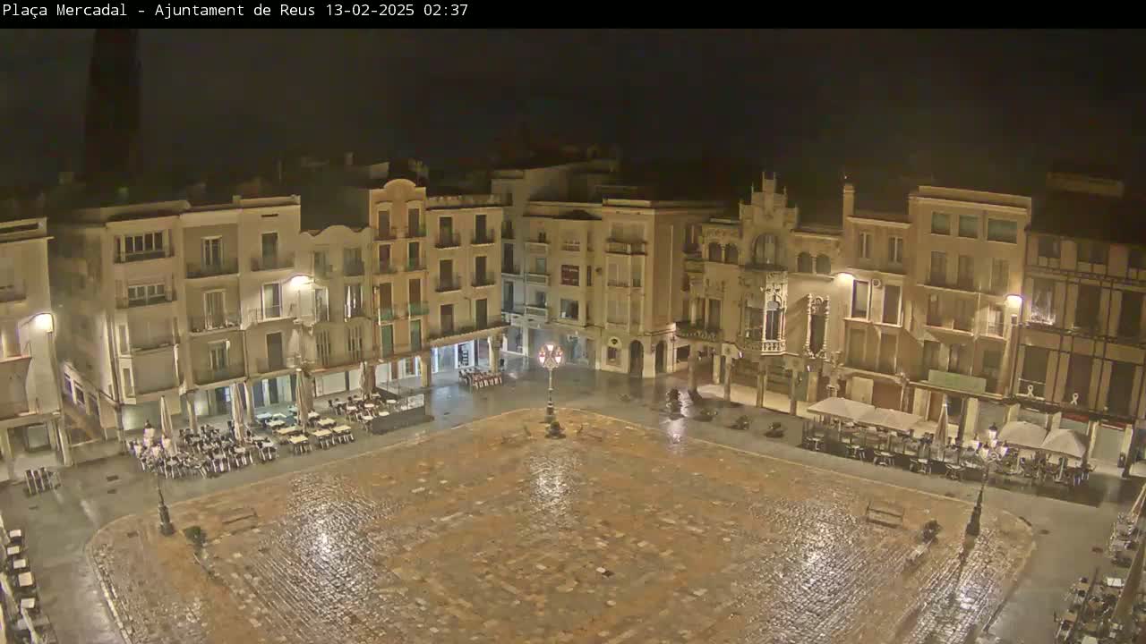 A rain-slicked, cobblestone town square at night, surrounded by multi-story buildings with outdoor seating areas.