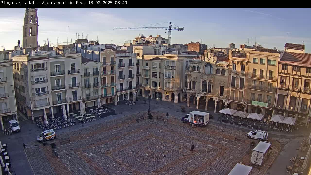 A large, mostly empty cobblestone town square is surrounded by several multi-story buildings under a clear, bright sky.