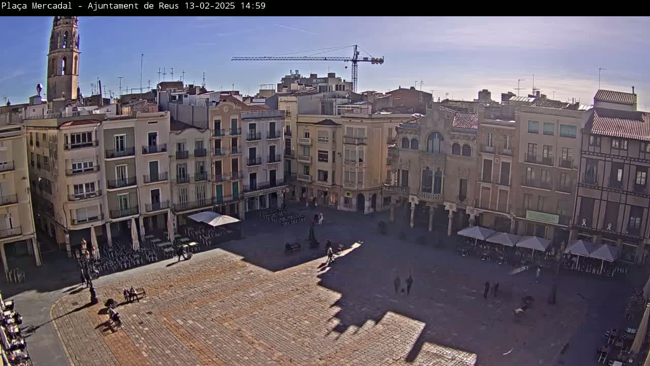 A sunny day reveals a large town square surrounded by multi-story buildings, with a few people scattered throughout and outdoor seating at cafes.